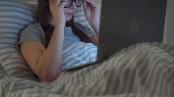 Женщина в очках работает на ноутбуке, лежа ночью в постели. Мобильная зависимость или бессонница . — стоковое видео
