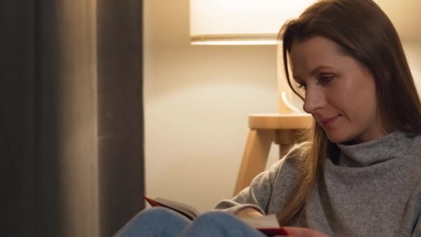 Vrouw liggend op de bank in een gezellige kamer en leesboek in de avond door het licht van een lamp — Stockvideo