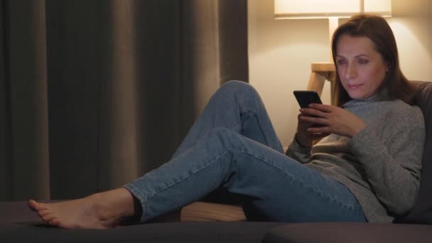 Smiling woman lying on the couch in a cozy room and using smartphone for surfing internet in the evening. Relaxation and lifestyle technology. — Stock Video