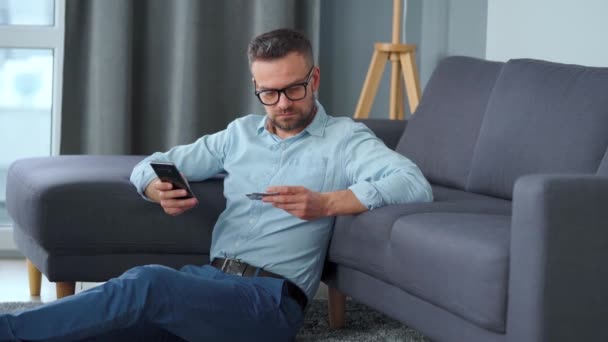 Hombre barbudo en gafas se sienta en el suelo cerca del sofá y hace el pago en línea con una tarjeta de crédito y un teléfono inteligente. Compras en línea, tienda en el sofá, tecnología de estilo de vida — Vídeos de Stock