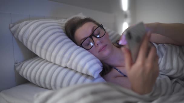 Mulher de óculos usando smartphone enquanto deitada na cama. Ela esfrega os olhos, porque está cansada e sonolenta. Conceito de aumento do estresse e fadiga . — Vídeo de Stock