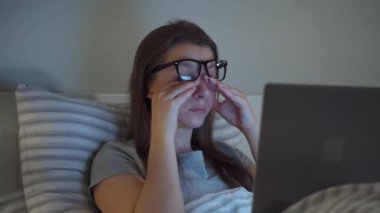 Woman in glasses working on laptop while lying in bed at night. She rubs her eyes, because she is tired and sleepy. Concept of increased stress and fatigue.
