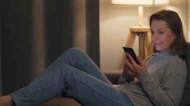 Sıcak bir odada kanepede uzanmış gülümseyen bir kadın ve akşamları internette sörf yapmak için akıllı telefon kullanıyor. Gördüğü için çok mutlu. Rahatlama ve yaşam tarzı teknolojisi.