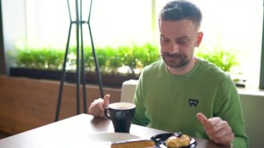 Bıyıklı sakallı adam çikolatalı ekler yiyor ve kafede kahve içiyor.