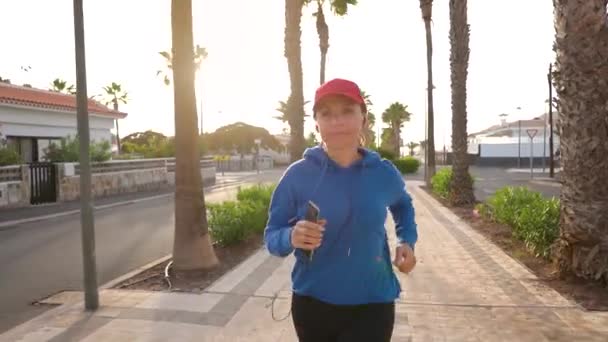 Woman with headphones and smartphone runs down the street along the palm avenue at sunset. Healthy active lifestyle — ストック動画