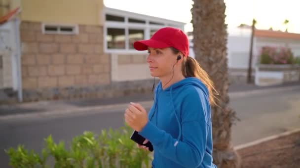 Kulaklıklı ve akıllı telefonlu bir kadın gün batımında Palm Avenue boyunca koşuyor. Sağlıklı aktif yaşam tarzı. Ağır çekim. Yan görünüm — Stok video