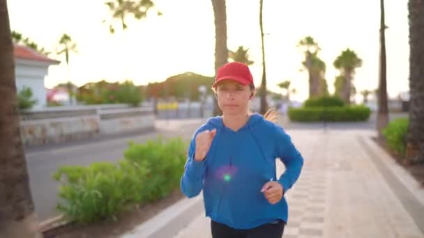 日落时，戴着耳机的女人沿着棕榈街跑着。健康积极的生活方式 — 图库视频影像