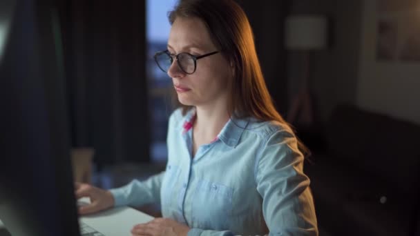 Een vrouw met een bril kijkt op de monitor en surft op internet. Het beeldscherm wordt gereflecteerd in de bril. Werk 's nachts. Binnenlandse Zaken. Afstandswerk — Stockvideo