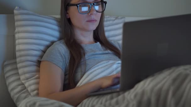 Жінка в окулярах працює на ноутбуці, лежачи в ліжку вночі. Інтернет-залежність або концепція безсоння . — стокове відео