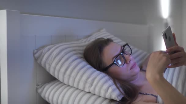 Femme en lunettes utilisant un smartphone alors qu'elle est couchée au lit. Elle se frotte les yeux, parce qu'elle est fatiguée et endormie. Concept de stress et de fatigue accrus . — Video