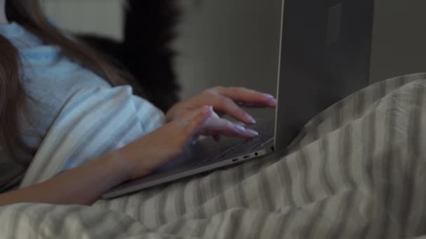 Woman typing on laptop while lying in bed at night. Black cat on the background. Internet addict or insomnia concept. — Stock Video