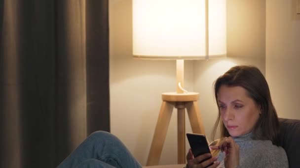 Mujer tumbada en el sofá en una habitación acogedora y el uso de teléfono inteligente para navegar por Internet en la noche. Tecnología de relajación y estilo de vida . — Vídeo de stock