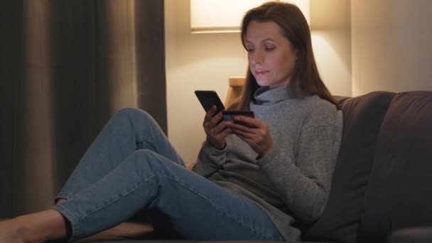 Женщина лежит на диване в уютной комнате и совершает онлайн-покупки с помощью кредитной карты и смартфона. Интернет-магазины, технологии образа жизни . — стоковое видео