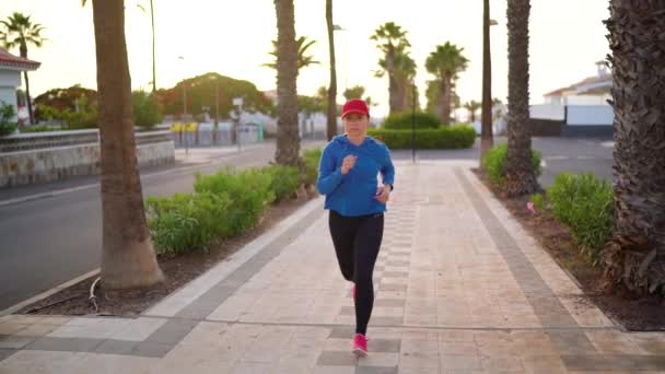 Γυναίκα με ακουστικά στ 'αυτιά τρέχει στο δρόμο κατά μήκος της λεωφόρου Παλμ το ηλιοβασίλεμα. Υγιής ενεργός τρόπος ζωής. Αργή κίνηση — Αρχείο Βίντεο