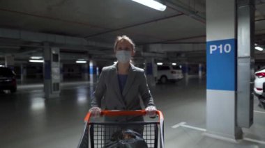 Tıbbi maskeli bir kadın bir market arabasıyla yeraltı otoparkından geçiyor. Covid-19 Coronavirus Pademia sırasında satın alma