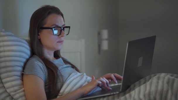 Gözlüklü kadın geceleri yatakta yatarken dizüstü bilgisayarda çalışıyor. İnternet bağımlısı veya uykusuzluk konsepti. — Stok video