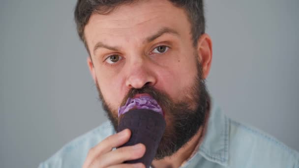 Close-up de um homem barbudo comendo sorvete roxo em uma xícara de waffle preto em um fundo de parede cinza — Vídeo de Stock