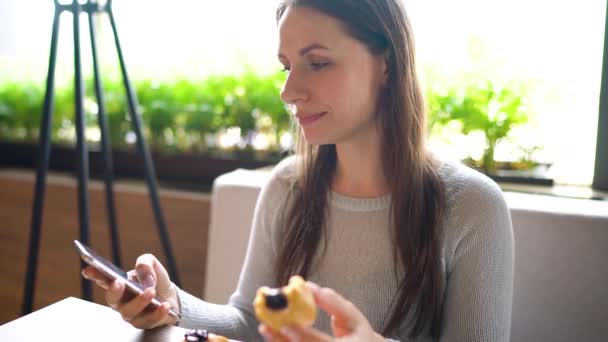 Vrouw die eclairs eet in een café en een smartphone gebruikt — Stockvideo