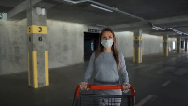 Eine Frau mit medizinischer Maske geht mit einem Einkaufswagen durch eine Tiefgarage. Kauf während Covid-19 Coronavirus-Pademie — Stockvideo