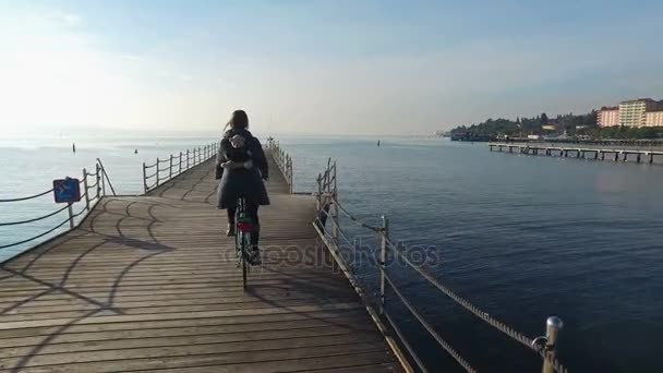 4К. Счастливая девушка катается на велосипеде по синему морю на пирсе в солнечный день. Время отпуска — стоковое видео