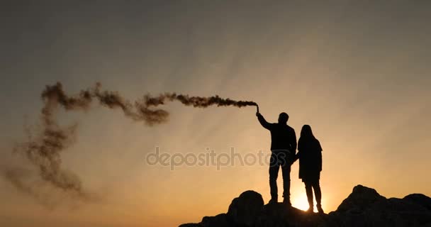 Dos amantes sostienen una bomba de humo en sus manos, en el fondo de una puesta de sol, firman el SOS — Vídeo de stock