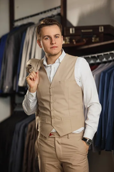 Empresario en chaleco clásico contra hilera de trajes en tienda. Un hombre joven y elegante con una chaqueta de tela negra. Está en la sala de exposición, probándose ropa, posando. Publicidad foto — Foto de Stock