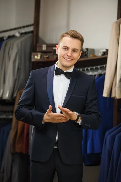 Empresário em colete clássico contra fileira de ternos na loja. Um homem jovem e elegante com um casaco de pano preto. Está no showroom, experimentando roupas, posando. Publicidade foto — Fotografia de Stock