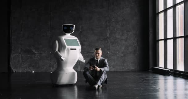 Επιχειρηματίας χρησιμοποιεί ένα smartphone, ενώ κάθεται στο πάτωμα δίπλα ένα ρομπότ. Σύγχρονη ρομποτικές τεχνολογίες. Αυτόνομο ρομπότ humanoid. — Αρχείο Βίντεο
