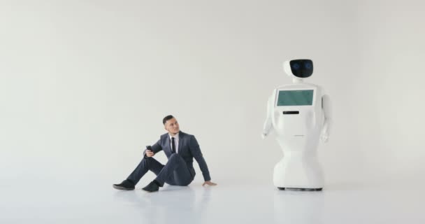 Zakenman maakt gebruik van een smartphone zittend op de grond naast een robot. Moderne Robotic technologieën. Autonome humanoidrobot. witte achtergrond — Stockvideo