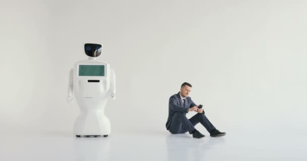 Uomo d'affari utilizza uno smartphone mentre seduto sul pavimento accanto a un robot. Tecnologie robotiche moderne. Robot autonomo umanoide. sfondo bianco — Video Stock