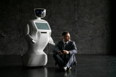 işadamı bir smartphone bir robot yanındaki yerde otururken kullanır. Modern robotik teknolojiler. Özerk insansı robot. çatı katı ofis