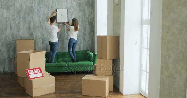 Jóvenes felices estudiantes de la pareja milenaria se mudan a su primera casa de nuevos propietarios. Fondo simple de apartamento urbano, enmarcan y deciden sobre la decoración de paredes, pintura y mapa — Vídeo de stock