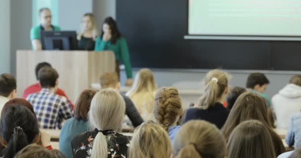 Grupo de estudiantes universitarios sentados en sus escritorios en el auditorio y escuchando mientras su profesor imparte una conferencia . — Vídeos de Stock