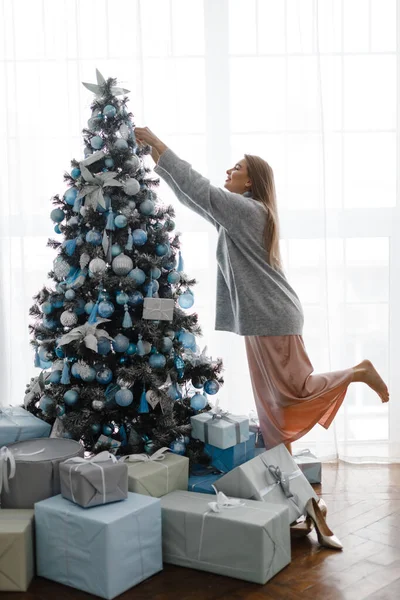 Hermosa chica decora un árbol de Navidad, de pie delante de la ventana . — Foto de Stock