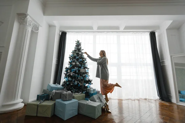 Vacker flicka dekorerar en julgran, står framför fönstret. — Stockfoto