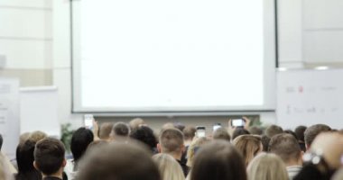 Geniş izleyici kitlesindeki izleyiciler sunum ve arka plan görüntüleriyle ekranı fotoğraflıyorlar.