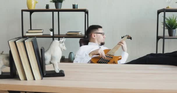 Νεαρός ονειροπόλος στο γραφείο παίζει κιθάρα καθισμένος στο τραπέζι με τα πόδια πάνω του. — Αρχείο Βίντεο