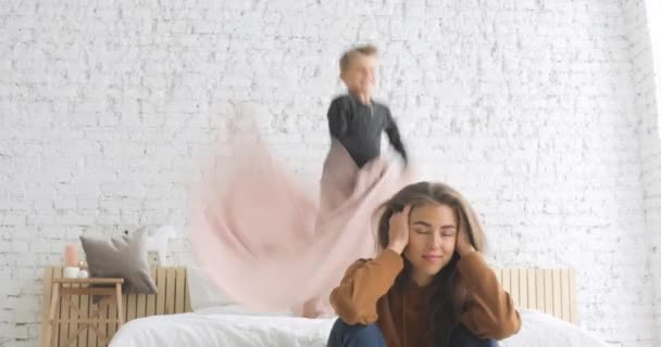 Гіперактивний дошкільний син хлопчик стрибає на ліжку, втомившись від шуму, який матуся прикриває вуха . — стокове відео