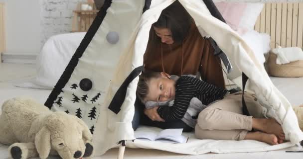 Moeder en zoon lezen samen een boek in tipi, jongen op vrouwenbenen. — Stockvideo