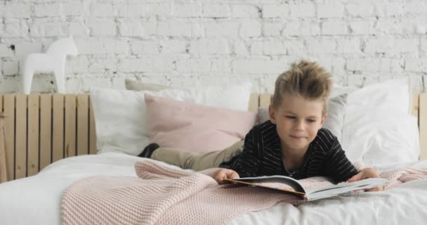 Junge liest ein Papierbuch auf dem Bett liegend im Schlafzimmer zu Hause, Frontansicht. — Stockvideo