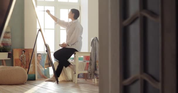 Pintora está pintando cuadro sentado cerca de la ventana en su estudio de taller moderno . — Vídeo de stock