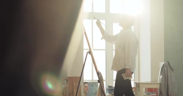 Paintress is drawing a picture on easel standing near the window, side view. — 비디오