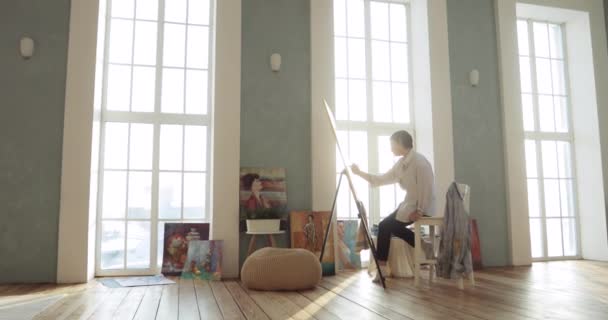 Pintura quadro pintor sentado no estúdio de arte perto de janelas, câmera zooms em . — Vídeo de Stock