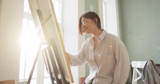 Mujer pintora está pintando cuadro utilizando cepillo sobre lienzo sentado cerca del caballete . — Vídeos de Stock