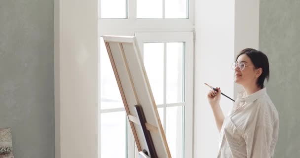 Γυναίκα ζωγράφος ζωγραφίζει εικόνα στο καβαλέτο στέκεται κοντά στο παράθυρο στο εργαστήριο. — Αρχείο Βίντεο