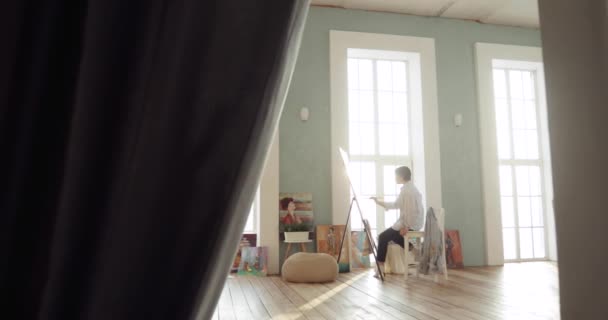 Schilderijenfoto zittend in kunststudio bij ramen, zicht van veraf. — Stockvideo