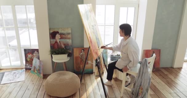 Paintress drawing picture on canvas on easel sitting in art studio near windows. — 비디오