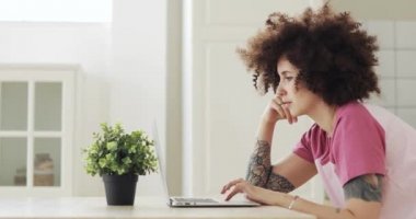 Young woman freelancer with curly hairs working on laptop at home, side view.