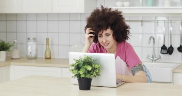 Joven mujer sonriente con pelos rizados charlando con amigos en el ordenador portátil en la cocina . — Vídeo de stock