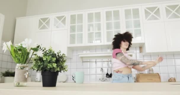 Щаслива молода жінка з кучерявим волоссям танцює і співає на кухні вдома . — стокове відео